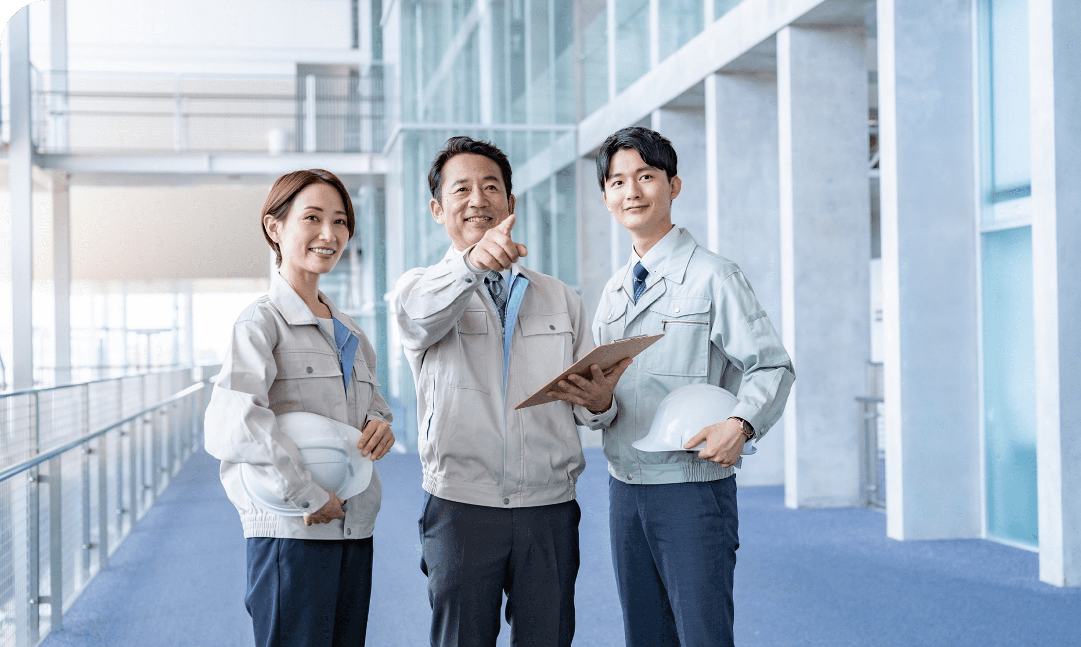 オフィス内で作業着を着た男性上司と女性と男性の部下が話している写真。上司はクリップボードを持ちながら遠くを指差し、部下たちは白いヘルメットを持っている。