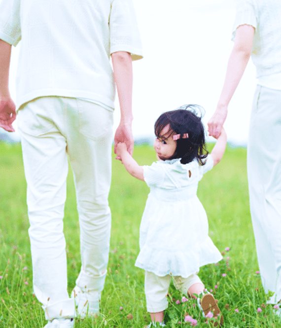 草原で白い洋服を着た両親と小さい女の子が手を繋いで歩き、女の子が振り向こうとしている写真