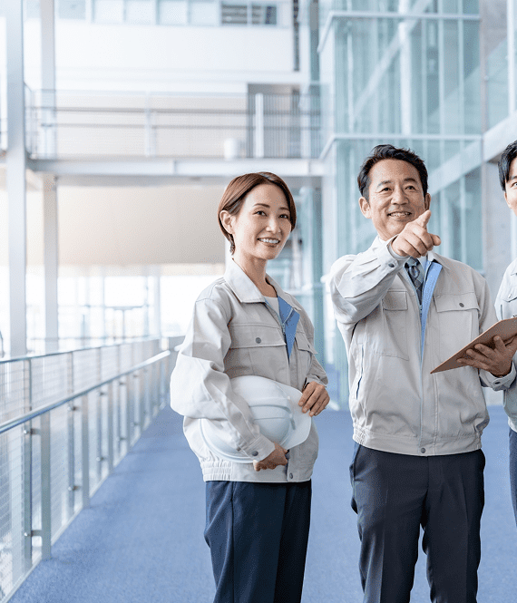 オフィス内で作業着を着た男性上司と女性と男性の部下が話している写真。上司はクリップボードを持ちながら遠くを指差し、部下たちは白いヘルメットを持っている。