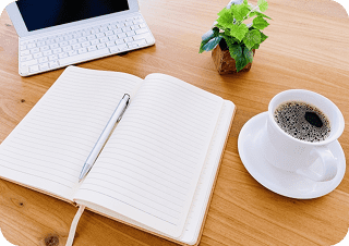 机にノートパソコン・ノートとボールペン・小さい観葉植物・コーヒーがある写真