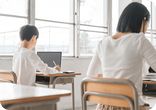 男の子と女の子が学校の教室の中で座ってタブレットを開いて勉強している後ろ姿の写真