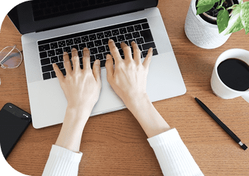 小さい観葉植物やコーヒー、眼鏡、携帯、鉛筆が置かれたデスクの上にあるノートパソコンでタイピングをしている女性の手が写った写真