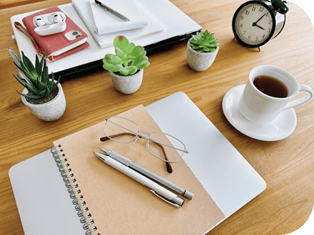 机の上にノートやペン・眼鏡が乗ったパソコンや同じくノートやペン・イヤホン・携帯が乗ったパソコン、コーヒーが入った白いカップや卓上時計、３つの小さい観葉植物がある写真