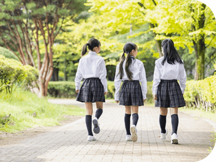 白いシャツにグレーのチェックスカート、紺のハイソックスの制服を着た3人の女の子が緑道を歩いている後ろ姿の写真