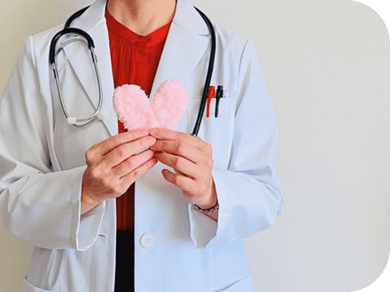 聴診器を首から下げ、胸ポケットに赤と黒のボールペンが入った白衣を赤いトップスの上に着た女性が、手にピンクのふわふわしたハートのオブジェを持っている写真