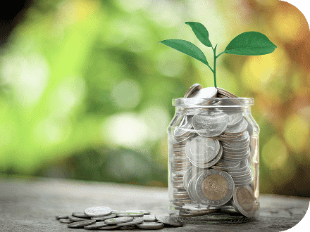 背景がぼやけ、机の上のお金が満杯に入っていて葉っぱのの芽が出てきている透明の瓶とそこから溢れたお金にピントが合った写真