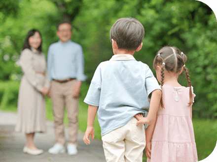 緑道に立っている後ろ姿の男の子と女の子から少し離れた向かいにピントがぼやけたおじいちゃんとおばあちゃんが立っていて、その孫たちとお互いに見つめ合っている写真